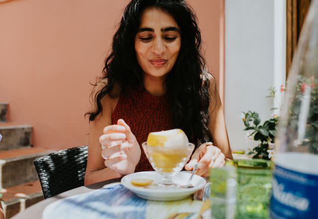 <p>Fiordaliso / Getty Images</p> A woman in a cafe uses a spoon to scoop lemon sorbet from a frozen lemon.