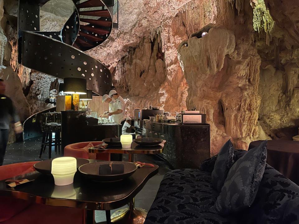 Image depicts the interior of a restaurant set in a cave. A spiral staircase leads down from the roof od the cave to the restaurant.