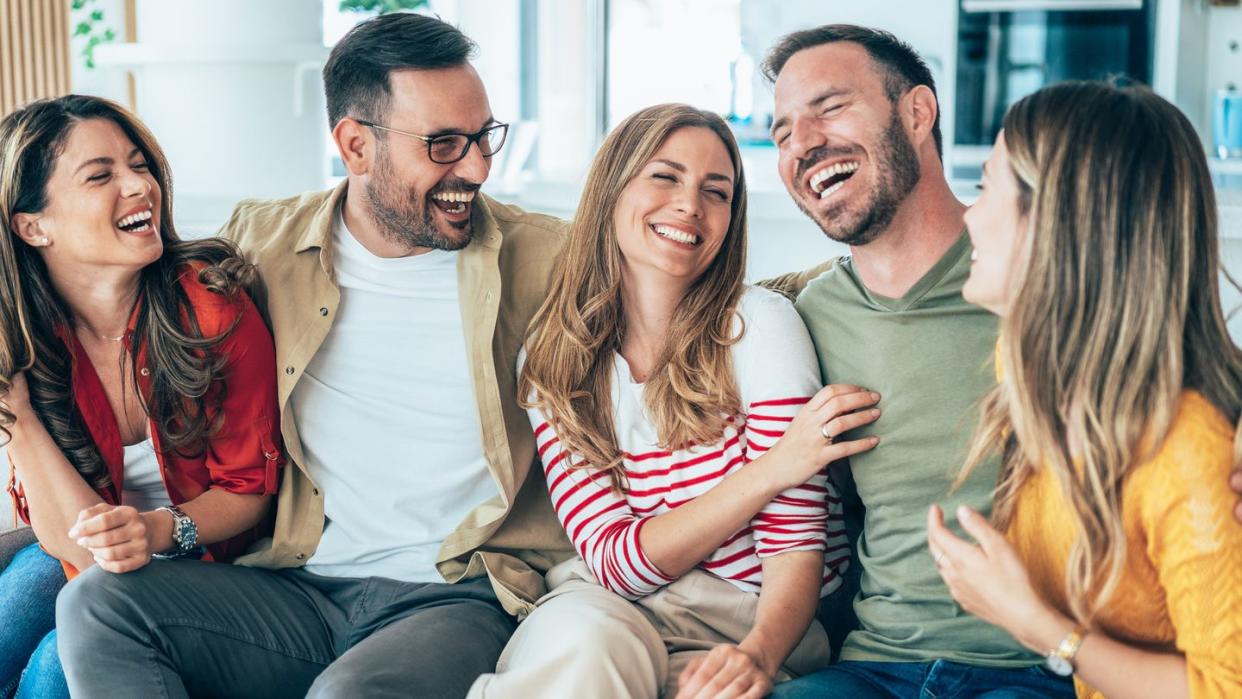 group of friends, two men and three women sitting on the sofa at home, talking and laughing they are wearing colorful, modern casual clothing