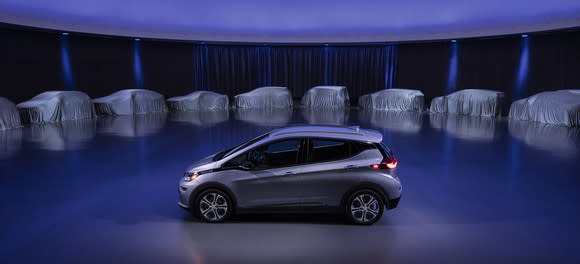 A silver Chevrolet Bolt EV on a stage, surrounded by 9 other vehicles hidden under white covers. GM said the covered vehicles represent some of its upcoming electric models.