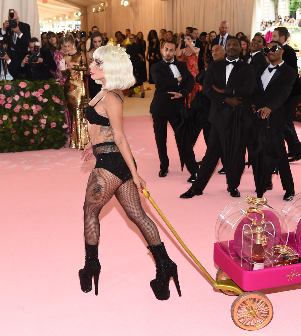 Lady Gaga walking on a pink carpet wearing a bold outfit with platform heels, pulling a cart with large perfume bottles. Photographers and guests in the background