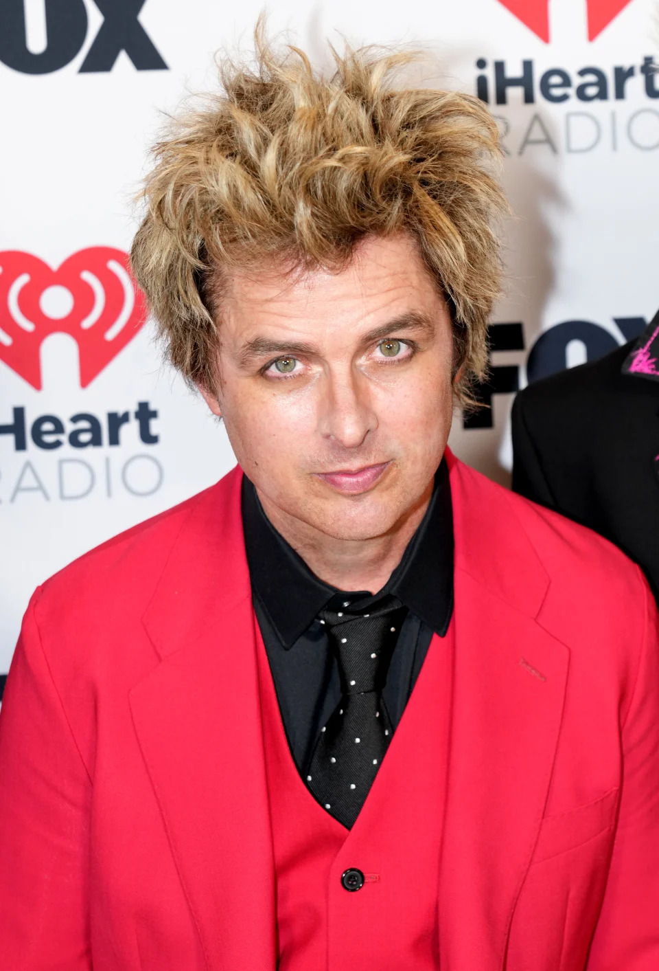 Billie Joe Armstrong at an iHeartRadio event, wearing a bright suit with a polka-dotted tie, and styled hair