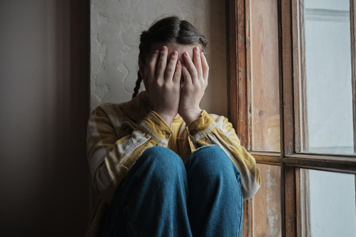 Upset depressed teenager covering face with hands and crying, sad worried teen girl sitting by window at home. Teenagers and mental health concept