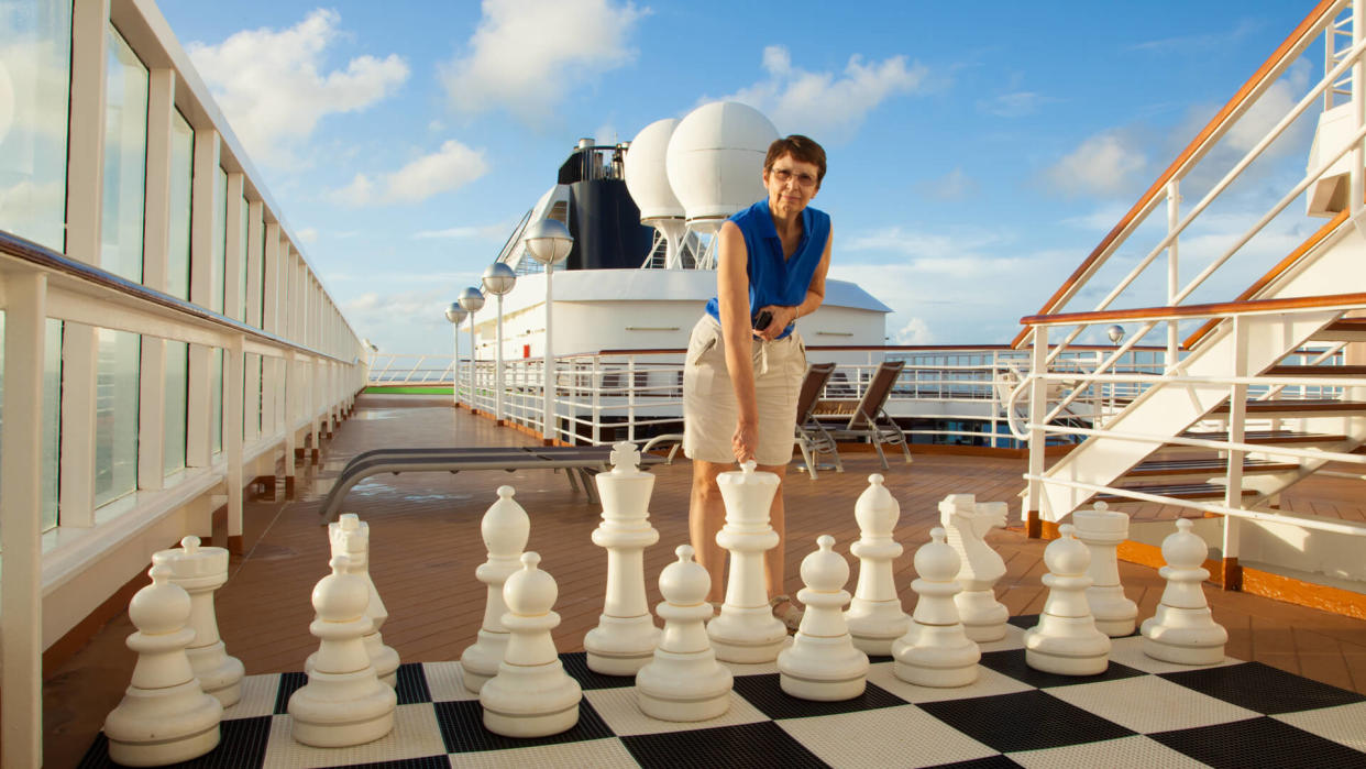 A mature, smiling woman prepares to move a large chess piece on a cruise ship.