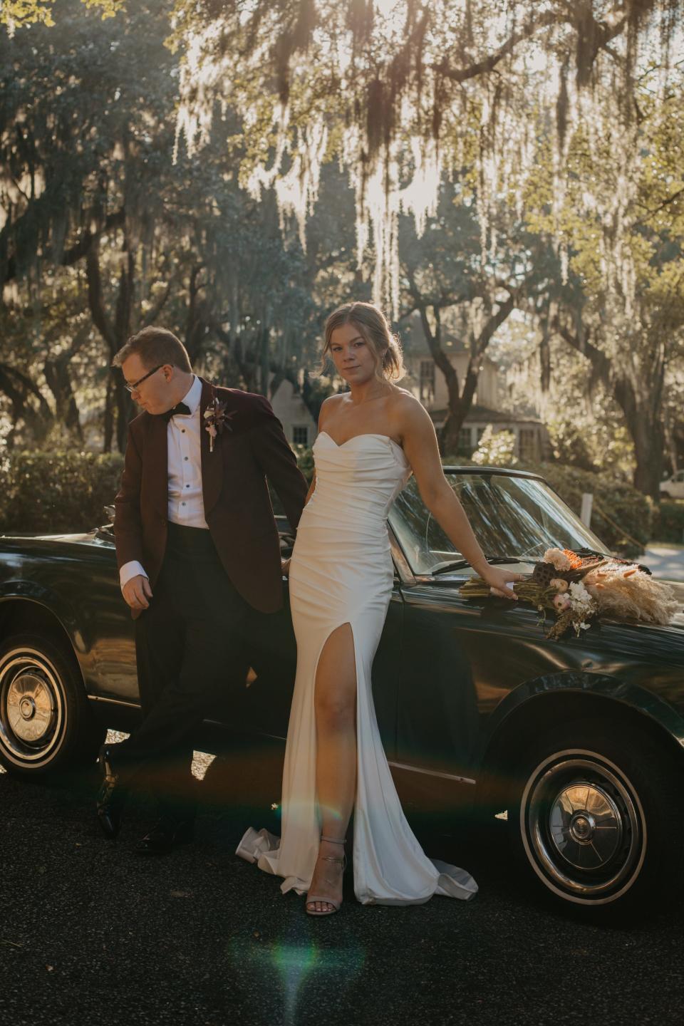 A bride and groom pose in front of a convertible in their wedding attire.