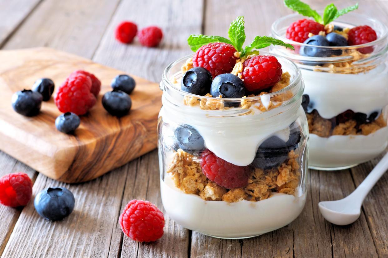 Two yogurt parfaits with strawberries, raspberries, and blueberries with one porcelain spoon on the right with an olivewood serving board on the left on a rustic wooden table, blurred in the background