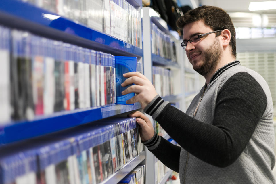 A man at a video game store with shelves full of games, pulling one out and looking at it with a half smile