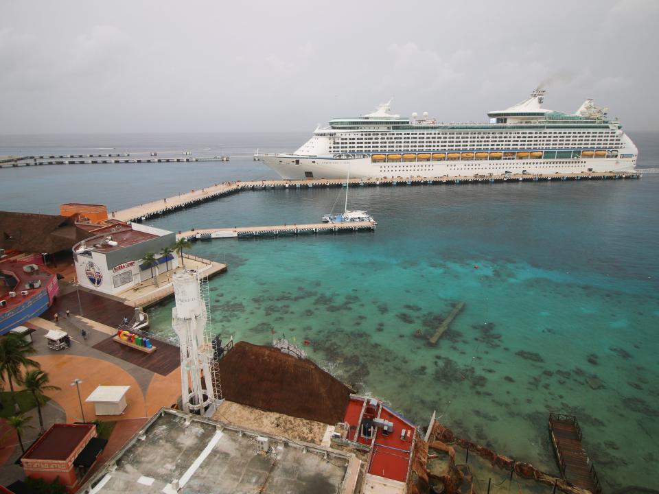 This image shows a cruise ship in the distance docked in Cozumel with buildings and palm trees and the beach in the foreground.