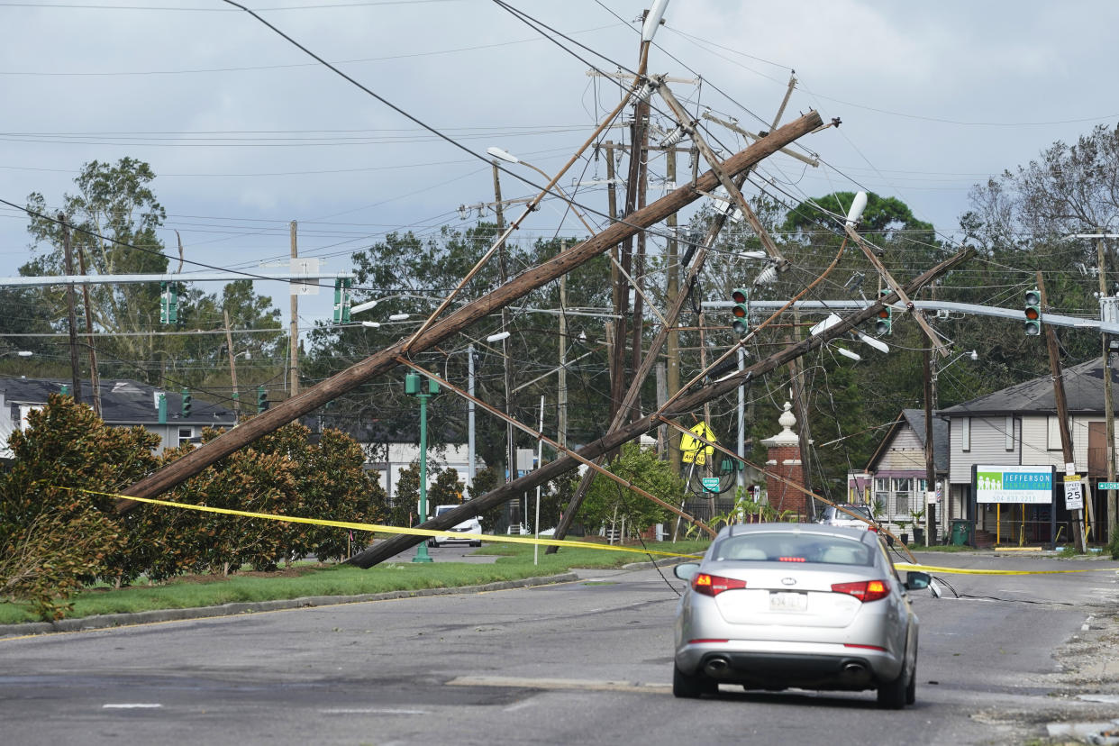 A car drives toward fallen utility poles wth yellow tape stretched across the street.