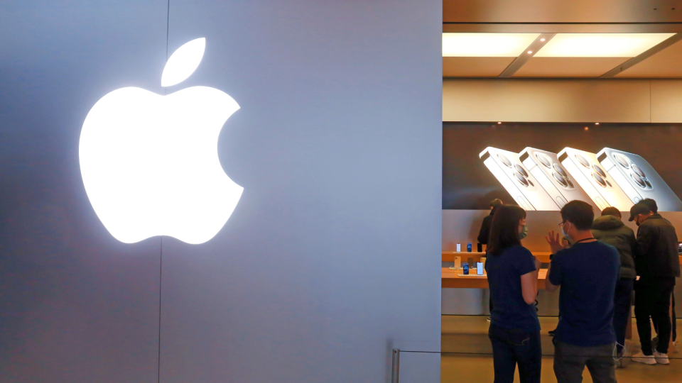 The exterior of an Apple store with the logo at the forefront and people using products in the background.