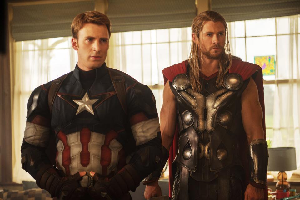 Chris Evans as Captain America stands beside Chris Hemsworth as Thor, both in superhero costumes, in a room with large windows