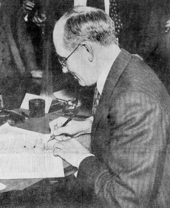 Michigan Gov. William Comstock is shown signing a document, sitting at a desk, wearing a suit.