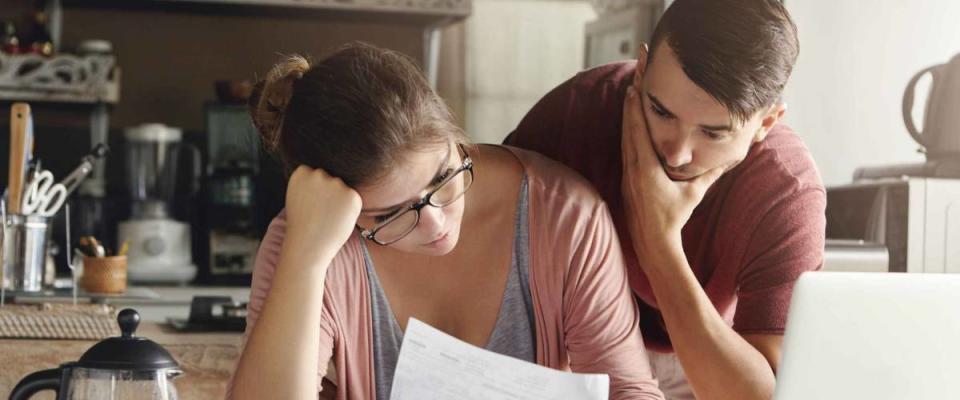 Young stressed Caucasian couple facing financials troubles, sitting at kitchen table with papers, calculator and laptop computer and reading document from bank, looking frustrated and unhappy