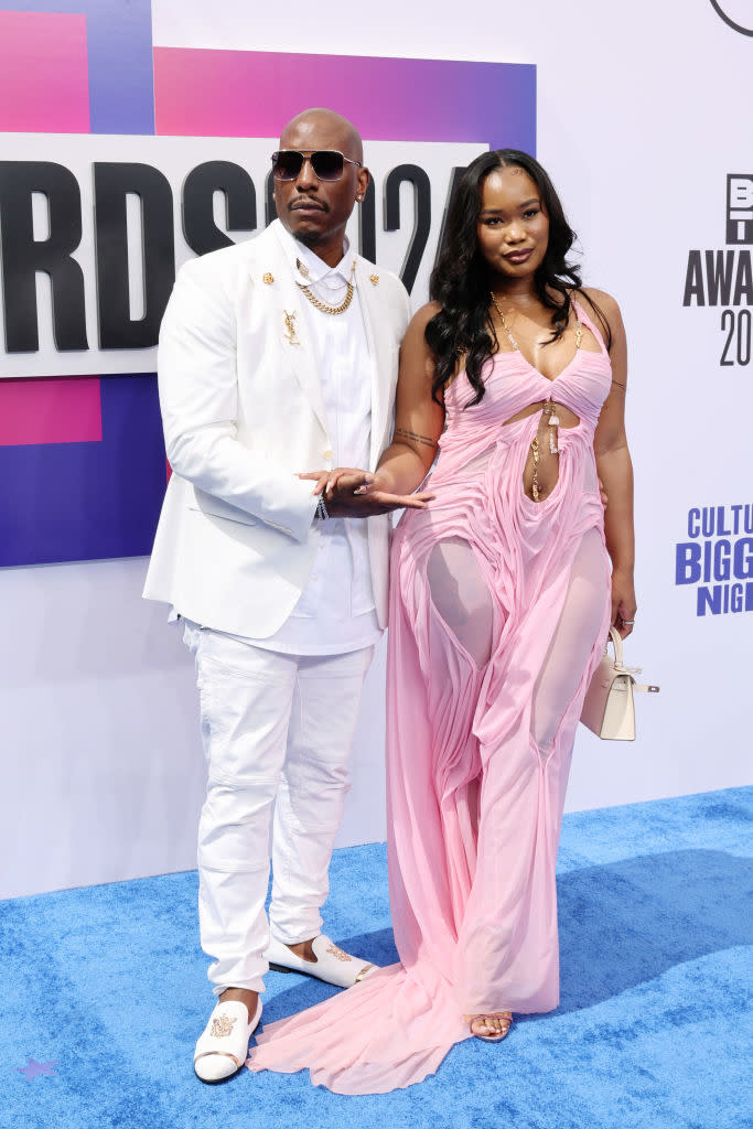 Tyrese Gibson and Zelie Timothy at an awards event. Tyrese wears a white suit, while Zelie wears a flowing, pink dress with cut-out sections.
