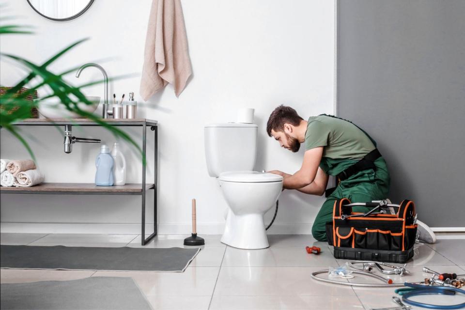 a man in green overalls fixes toilet plumbing in a large modern bathroom.