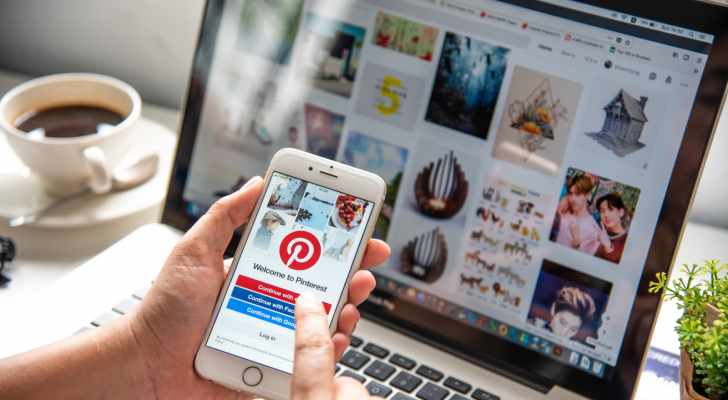 Hand holding Apple Iphone6 gold color with Pinterest app on the screen. In the background, a laptop is open to Pinterest. PINS stock.