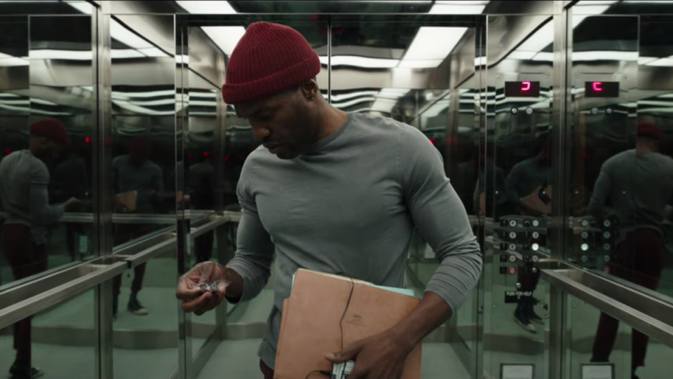 a photo of Yayha Abdul Mateen in Candyman horror film trailer wearing a grey shirt, red knit cap, and standing in an elevator with mirrors