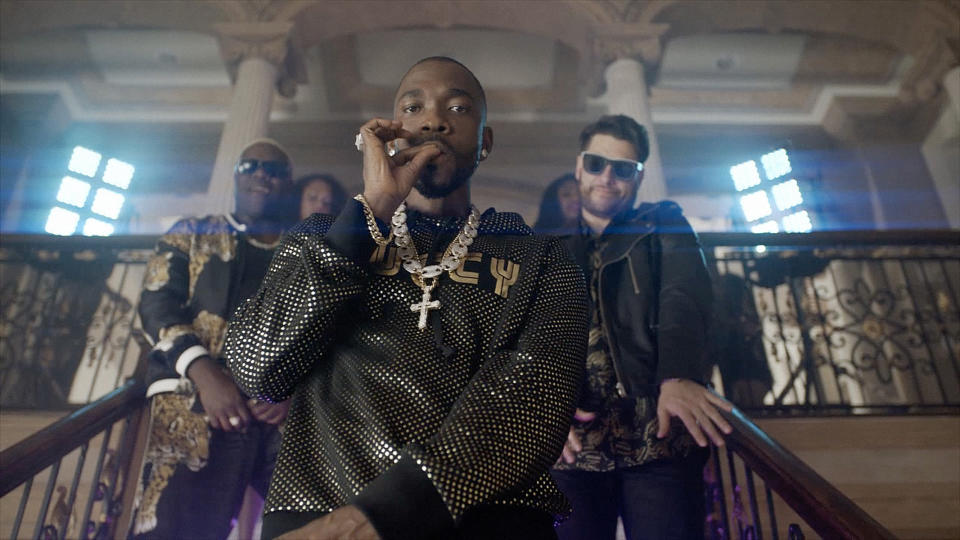 Three men on a staircase, one in a mesh top with chains, others in jackets, posed confidently