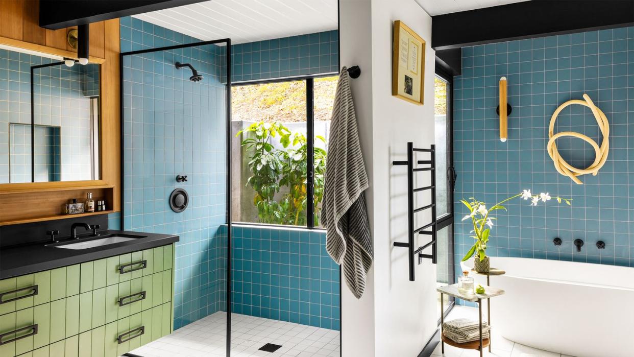 the primary bathroom has a walk in shower with aqua colored tiles, black shower and sink fixtures and countertop with green drawers below and mirror above, deep white bathtub with a sconce and wall sculpture above it