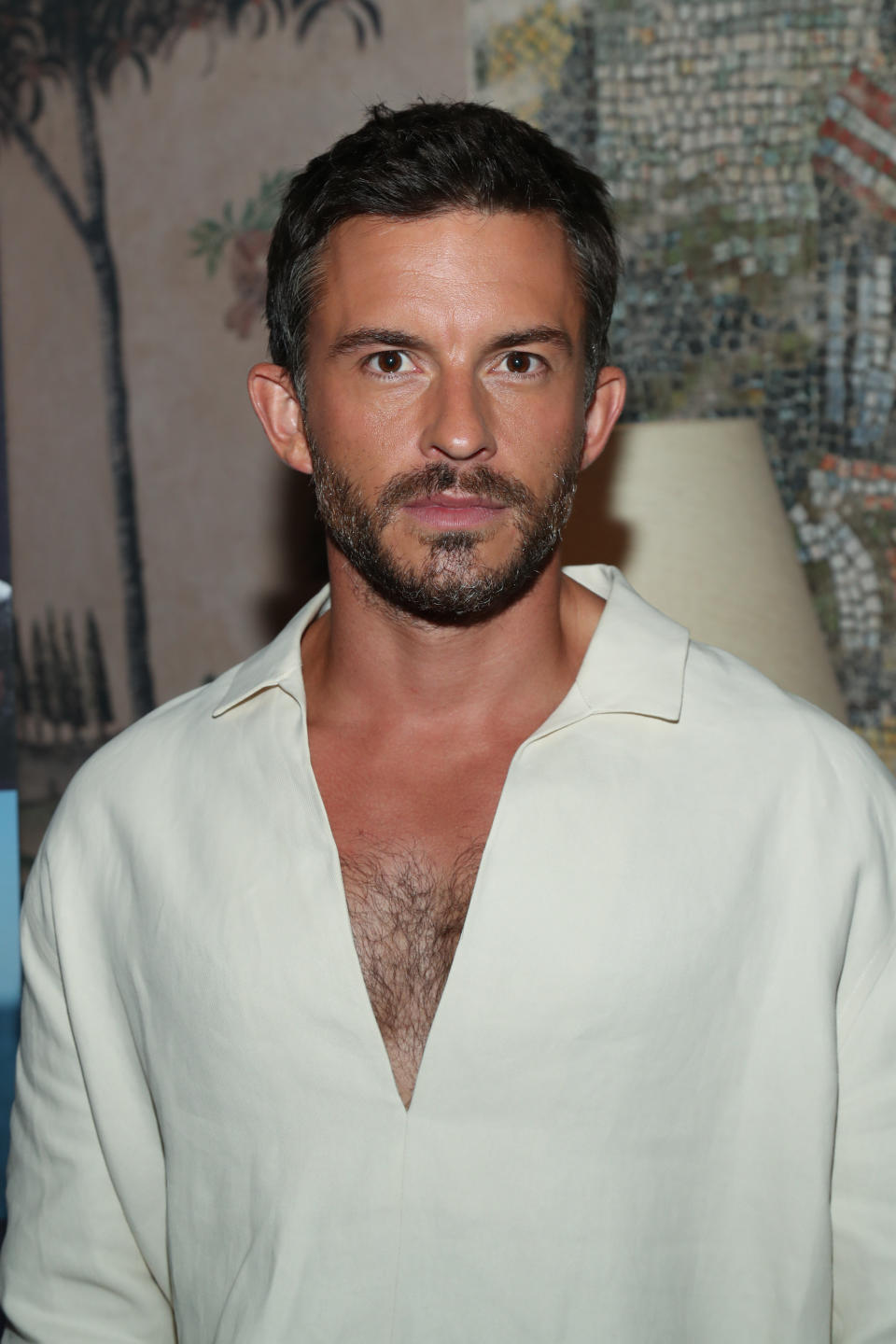 Jonathan Bailey with short hair and a trimmed beard, wearing a deep V-neck white shirt, stands for a photo