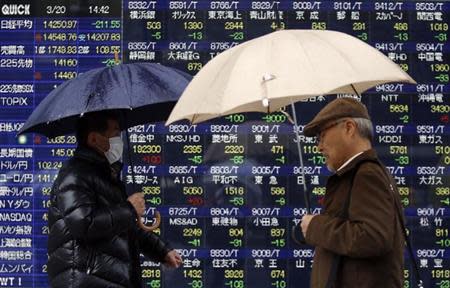 Pedestrians holding umbrellas walk past an electronic board showing stock prices outside a brokerage in Tokyo March 20, 2014. REUTERS/Yuya Shino
