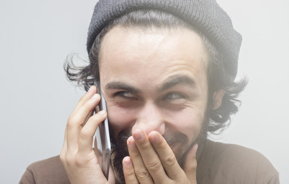 Portrait of young man using phone looking away with smirking facial expression. There is a deceitful human expression on his face. Bad man standing in front of gray background. There is a gray glowing light around the man. Smiling young man covering his mouth with his hand. Horizontal composition. Image taken in studio and developed from RAW format. Close-up portrait.