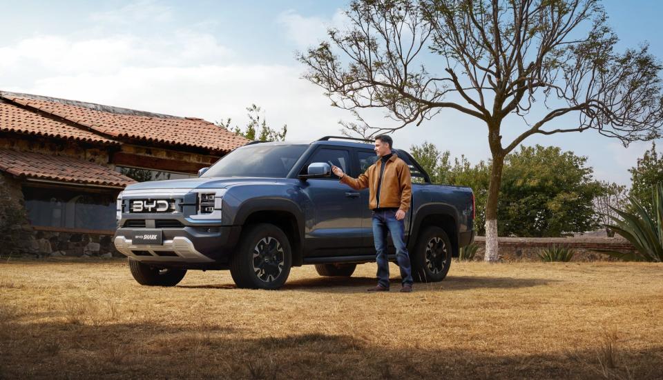 A man is standing next to a blue BYD Shark hybrid pickup truck parked on the grass in front of a house.