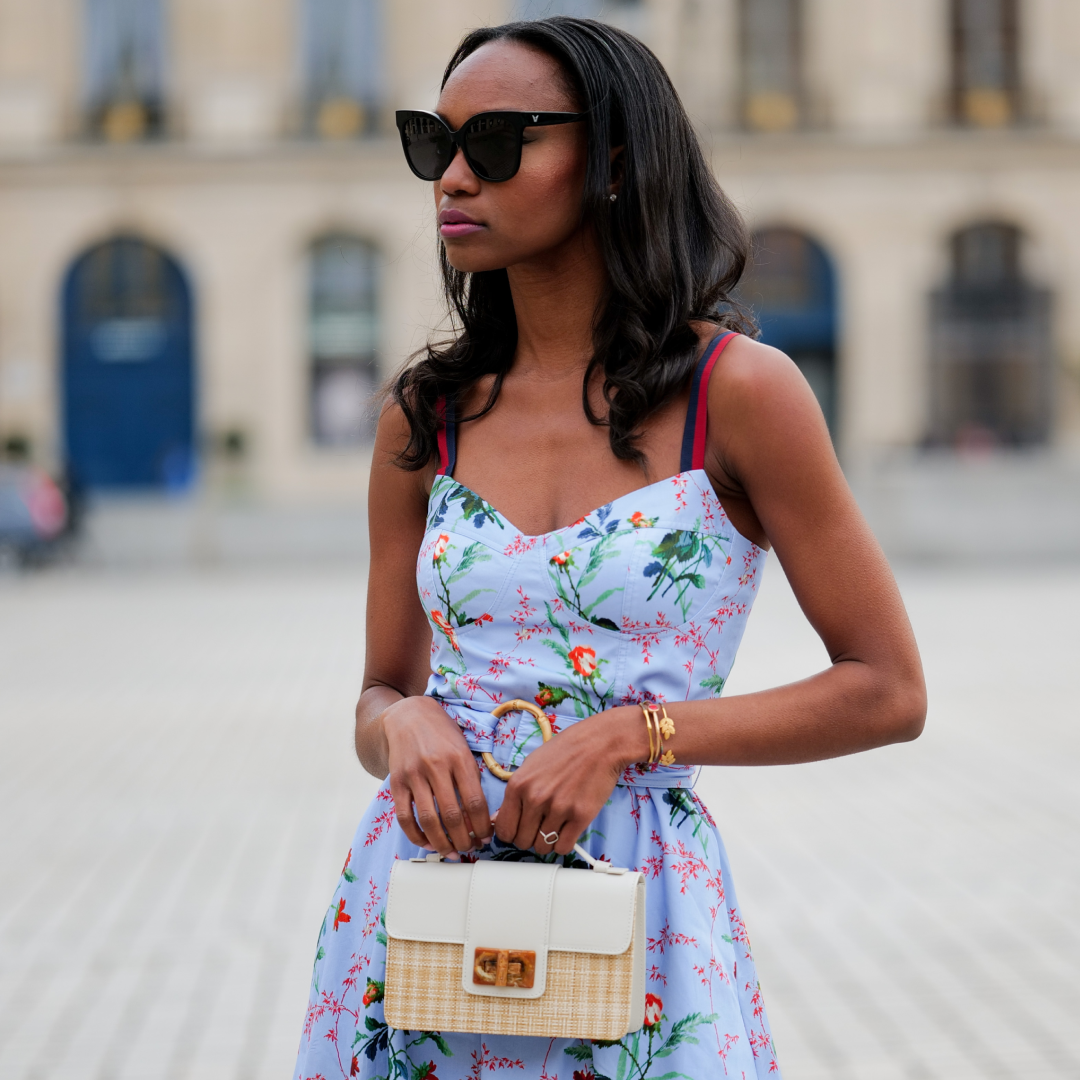  Milie Joseph wears black large sunglasses, crystal earrings, a pale blue with red / green / navy blue / beige flower print pattern heart neck / tank-top / belted / midi dress, a beige wickers and white shiny leather bi-material handbag, during a street style fashion photo session, on April 29, 2023 in Paris, France. . 