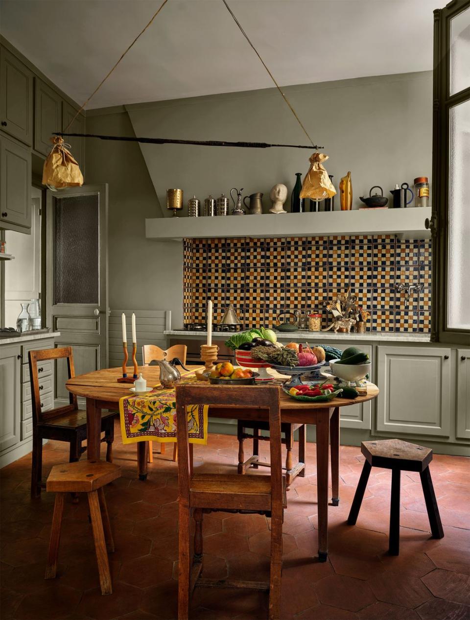 a kitchen has muted sage colored walls, a checkerboard tile backsplash and a shelf above it with vessels, round wood table with rustic chairs and stools, a triangle pendant, terra cotta floor