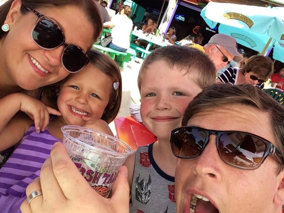 Selfie photo of Terri Peters, her husband, daughter, and son. Terri wears sunglasses and green studded earrings. To her right, her daughter wears a purple striped dress, a heart hair clip, and has blue eyes. Her brother stands to her right and has closely cropped dirty blonde hair, hazel eyes, and wears a grey Transformers tank top. Terri's husband wears sunglasses and has brown, short hair, and wears sunglasses. His mouth is open in faux surprise, posing.