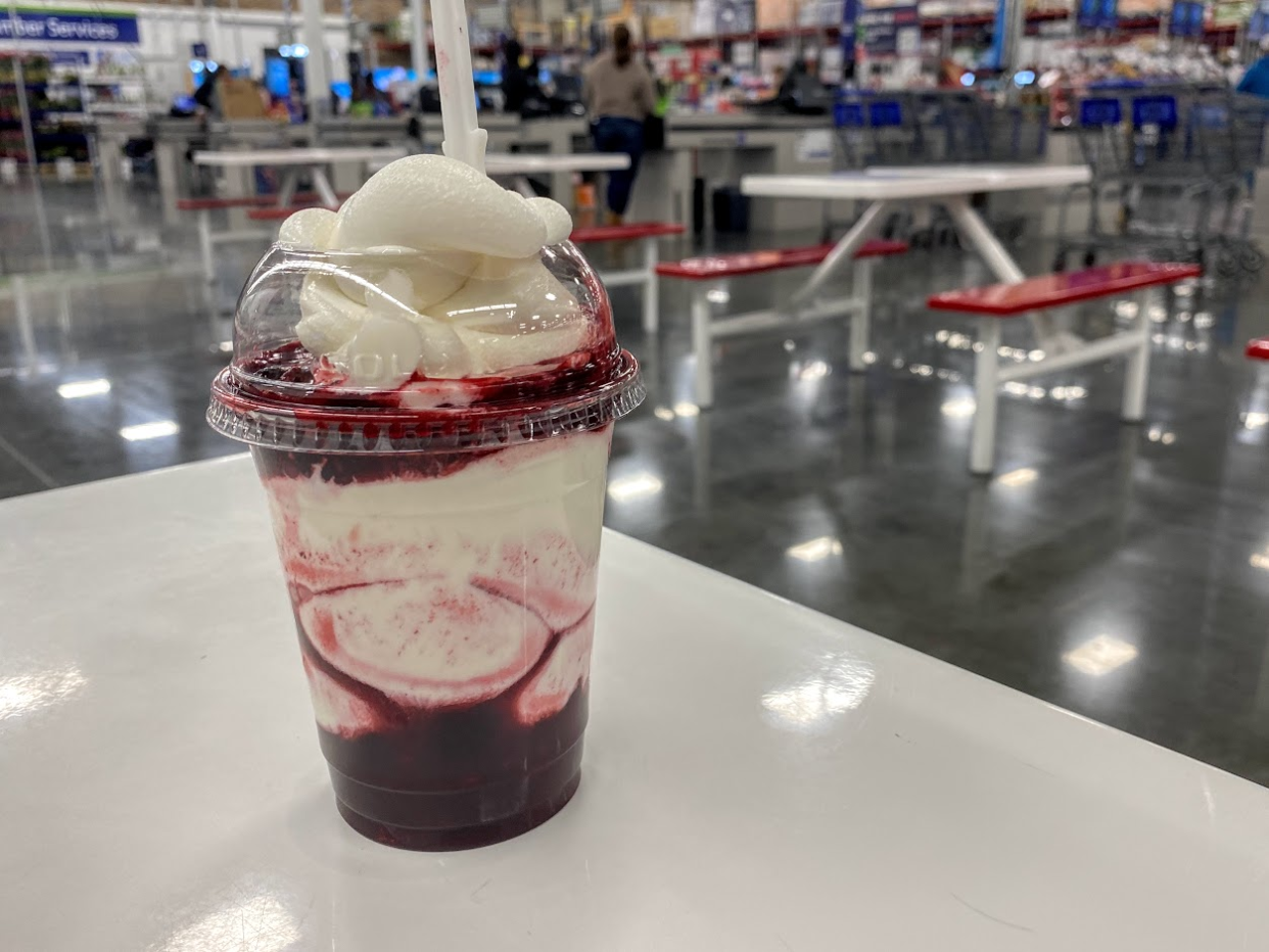 Berry sundae at Sam's Club cafe on a white table, grey concrete floor, cashiers, and warehouse blurred in the background