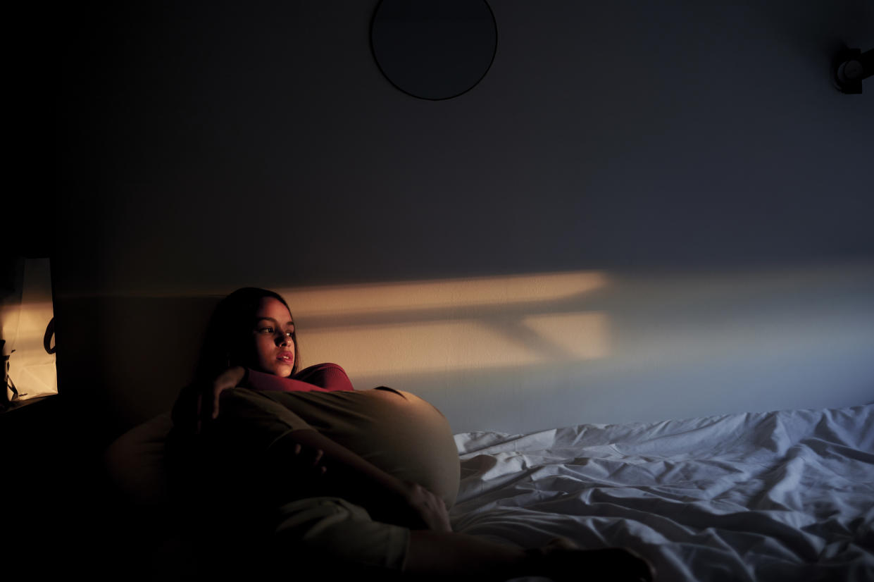 A woman sitting on a bed in a dark room looking out the window.