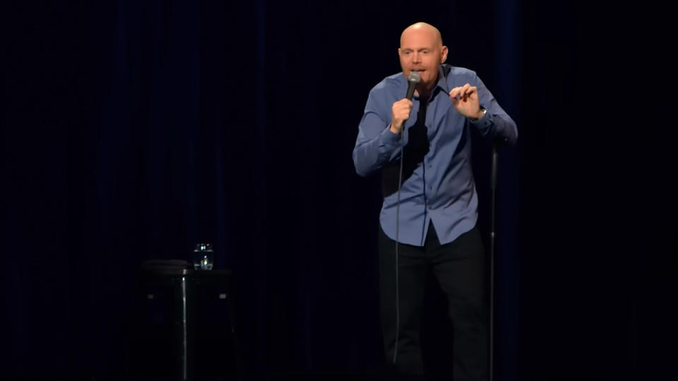 Bill Burr in a blue shirt, leaning on his microphone stand.
