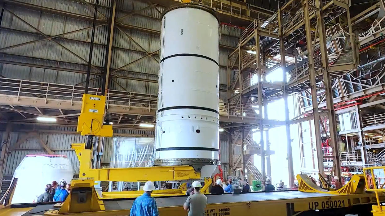  A large white cylinder is held vertical inside a warehouse building with hardhat-wearing folk standing about. 