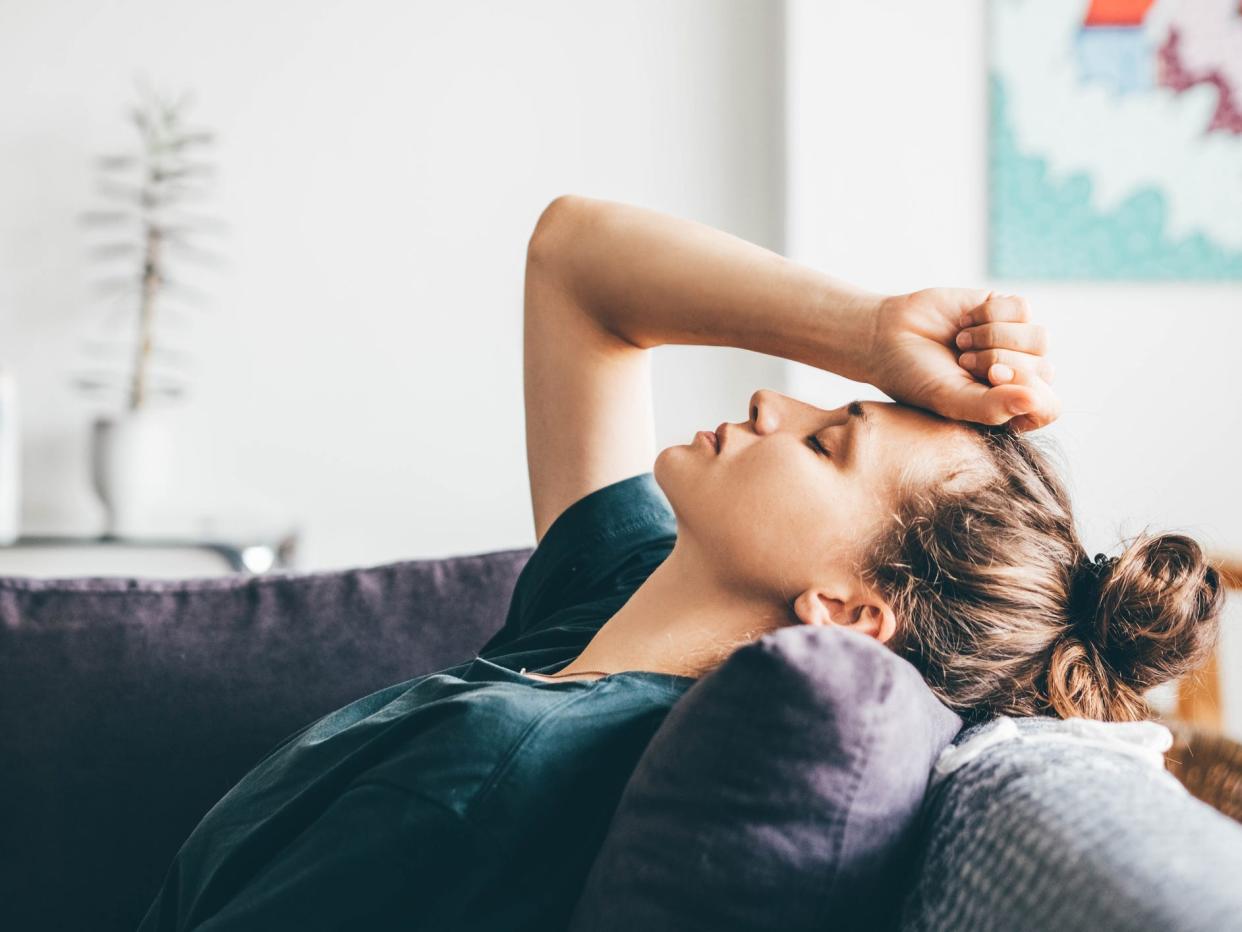 A woman with dirty blonde hair in a messy bun, sitting on a grey sofa with her eyes closed, leaning back against the pillow. Her right hand is in a fist on her forehead.