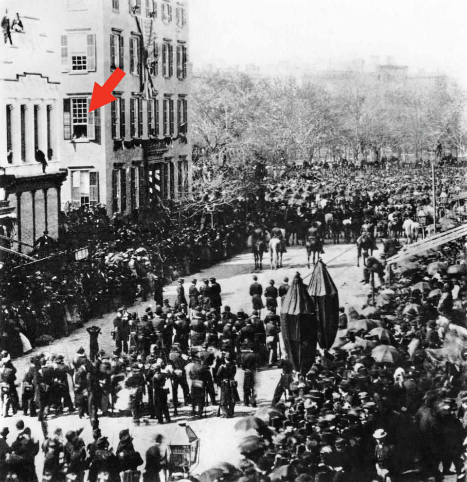 Historic photo of a large crowd gathered for a parade, possibly a military or political event, with buildings and trees in the background