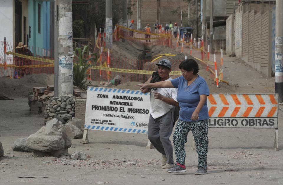 In this Feb. 12, 2020 photo, residents walk past signs that read in Spanish "Intangible archeological zone. Entrance prohibited," left, and "Danger. Works." where workers digging for a natural gas line ran into ancient bones and vessels from a previous Inca culture in the Puente Piedra neighborhood of Lima, Peru. About 300 archaeological finds, some 2,000 years old, have been reported over the past decade during the building of thousands of kilometers (miles) of natural gas pipelines in the capital. (AP Photo/Martin Mejia)