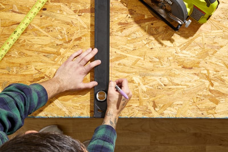 Overhead shot of a man holding a large level in place on a piece of plywood and drawing lines with a purple sharpie onto the wood.