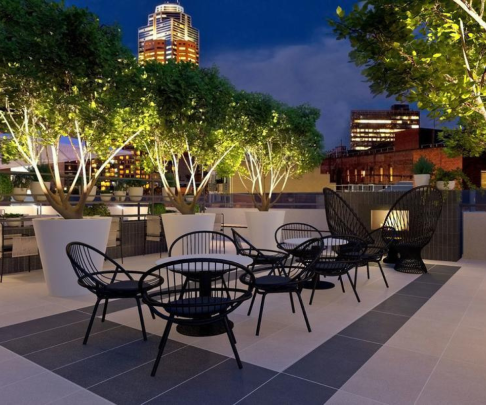 The rooftop terrace of Melbourne's Sheraton Hotel at dusk with a collection of black round outdoor chairs and trees in pot plants in a circle in the foreground with a lit up city scape in the background