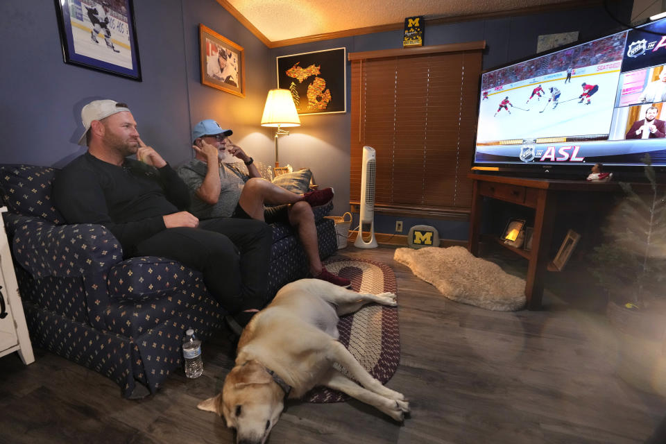 Bob Madden, center, and his son Jonathan, left, watch the Stanley Cup hockey final from a couch, Monday, June 10, 2024 in Genoa Township, Mich. Madden is deaf and he was enjoying the view with access to the commentary delivered in American Sign Language for the first time during a sporting event. The NHL became the first major sports league to offer play by play and analysis in ASL during a live broadcast. (AP Photo/Carlos Osorio)