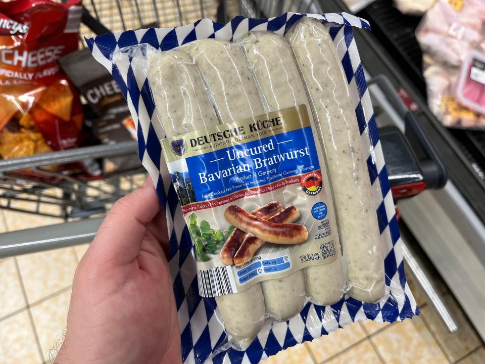 Eine Hand hält eine Packung mit vier ungepökelten bayerischen Bratwürsten der Aldi-Marke Deutsche Kuche. Auf der Verpackung ist ein blau-weißes bayerisches Muster zu sehen.