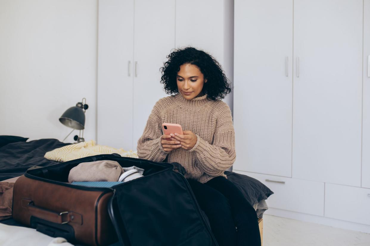 Beautiful woman packing her clothes into a suitcase and preparing for a journey while using her smartphone for online check in