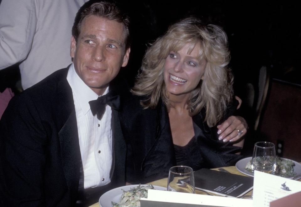 ryan o'neal and farah fawcett sit at a table and look to the left, he wears a black tuxedo, she wears a black outfit