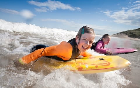 Two little girls are smiling for the camera while bodyboarding in the sea - Credit: DGLimages/Getty Images Contributor