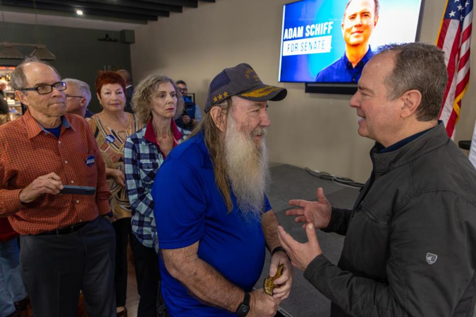 People stand by as Adam Schiff speaks with a man near a large screen showing his photo and reading "Adam Schiff for Senate"