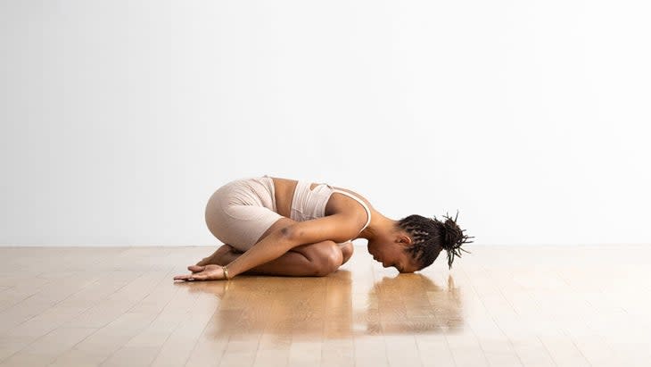 A Black woman wearing cream colored tights and top practices Child's Pose (Balasana). She is on a wood floor against a white backdrop.