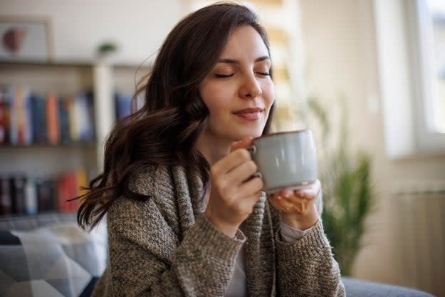 A woman enjoys a moment of relaxation while holding and savoring the aroma of a steaming cup of coffee, seated comfortably in a cozy room