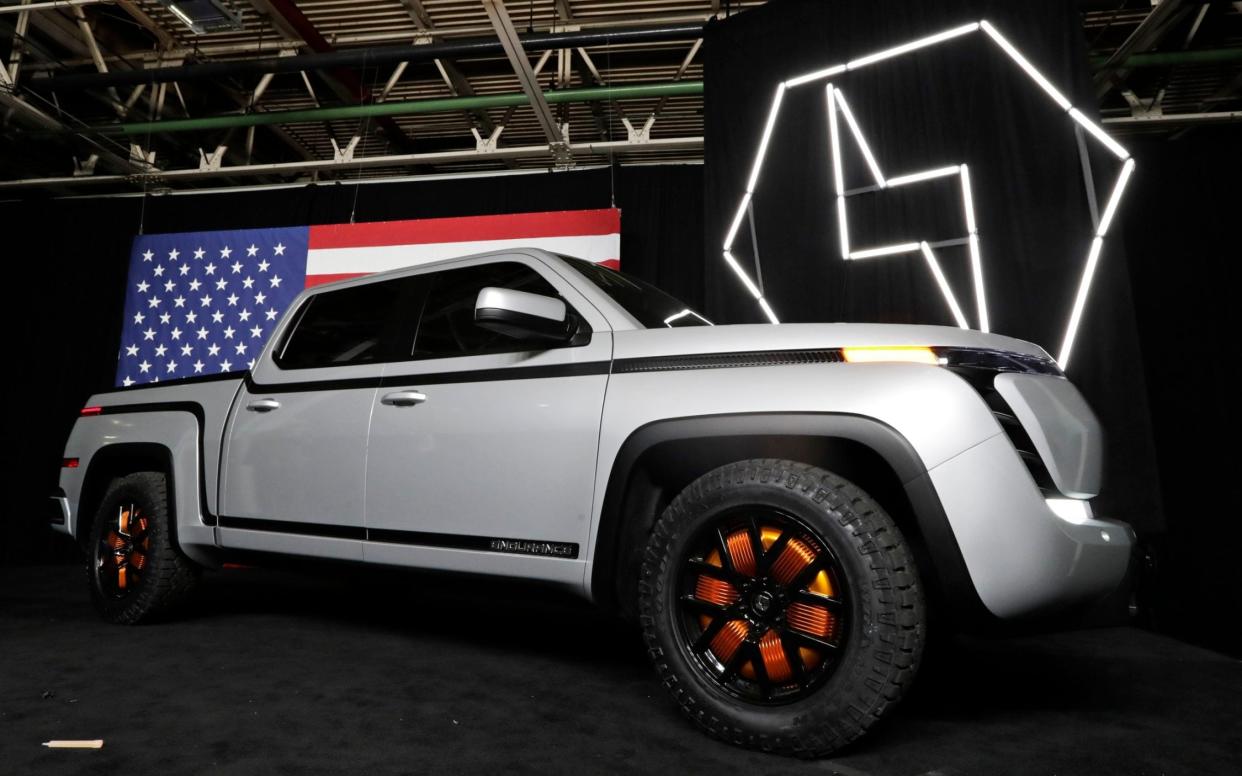 A bulky silver pick-up truck with curved contours and no front grille sits imposingly in front of a giant American flag and the lightning bolt logo of Lordstown Motors - Tony Dejak/AP