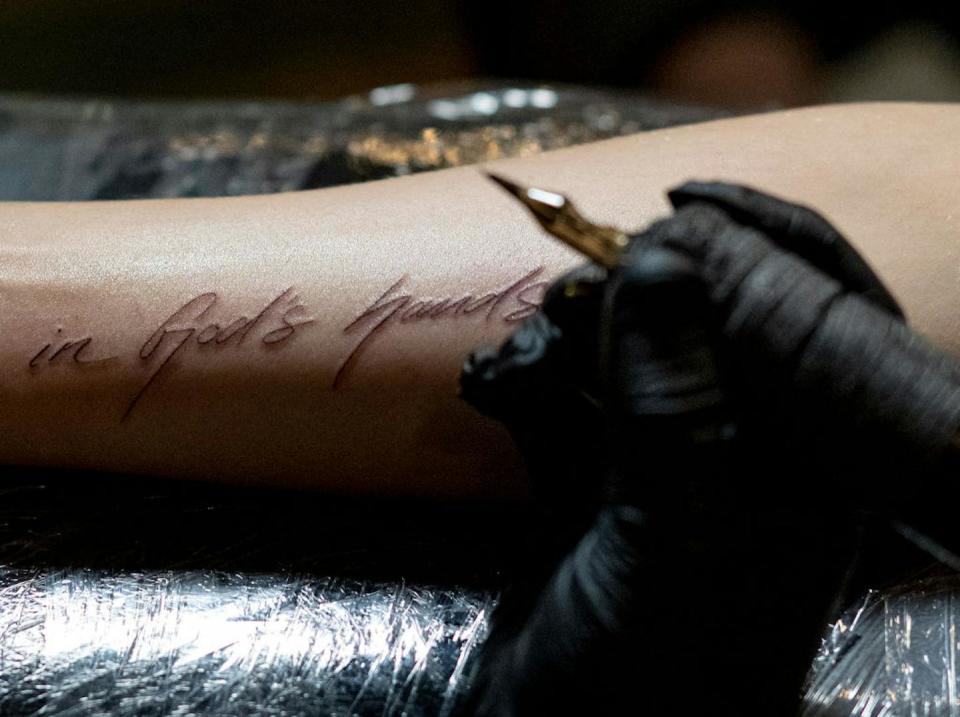 A hand in a black glove holds a golden tattoo needle over an arm inscribed 'in God's hands.'