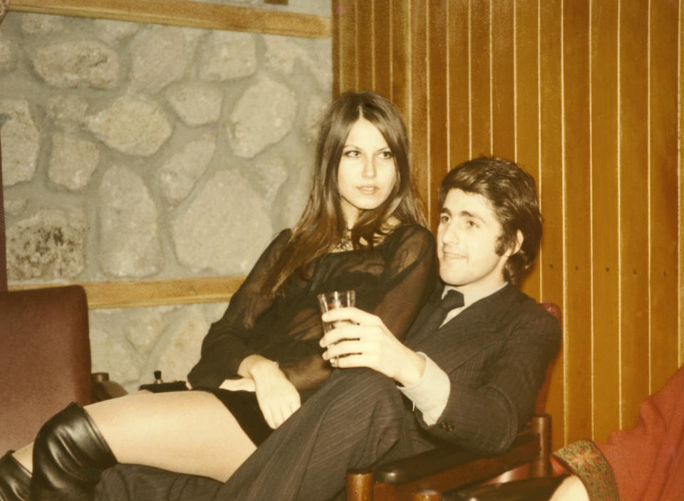 A woman in a short dress and knee-high boots sits on a man's lap. Both are holding drinks and looking off-camera